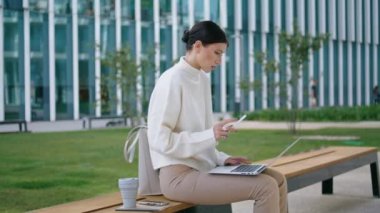 Serious smart woman working on laptop sitting at bench outdoors. Worried attractive businesswoman solving work problems remotely. Confident stylish lady browsing surfing internet outside near office.