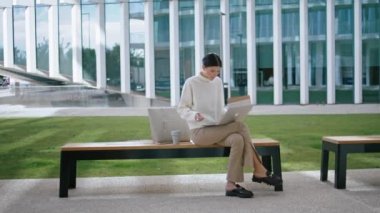 Smart young girl studying online using laptop on bench near city office. Focused pretty woman reading internet information holding paper sinopsis. Stylish busy businesswoman taking remote courses.