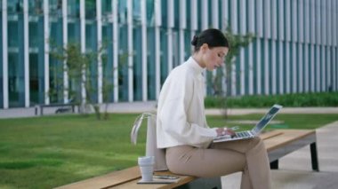 Smart business woman working remotely on laptop sitting at bench outdoors. Attractive focused lady typing report email messages on computer keyboard. Serious girl using notebook for freelance job.