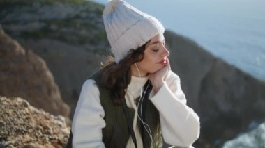 Gülümseyen kız dağın tepesinde kulaklık dinliyor. Huzurlu bir kadın okyanus kayalıkları manzarasında gözlerinizi kapatarak şarkıdan zevk alıyor. İyi tatiller. Uyum stresi olmayan yaşam tarzı