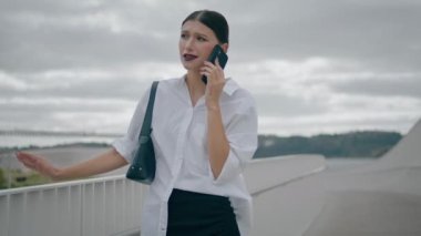 Young serious business woman dissatisfied phone conversation standing near railings cloudy day. Worried stylish lady having displeased telephone talk outdoors. Girl complaining at problems on mobile.