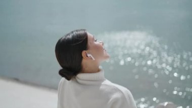 Carefree young woman putting face under sunlight sitting on waterfront alone closeup. Calm happy girl tourist relaxing near beautiful sparkling water in wireless earbuds. Cute brunette listening music