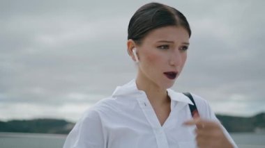 Portrait of elegant confident woman in wireless earbuds talking in front gray cloudy sky. Attractive successful business lady speaking phone using modern headset close up. Happy girl standing outdoors