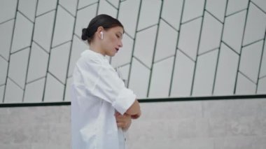 Thoughtful young girl secretary walking near modern office building close up. Serious attractive woman going to work holding paper documents on street. Lady freelancer in wireless earbuds enjoy stroll