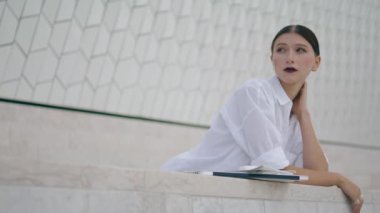 Calm attractive girl waiting meeting outdoors cloudy day. Confident business woman leaning on fence standing near modern building. Stylish ceo manager wearing white shirt relaxing on work break alone