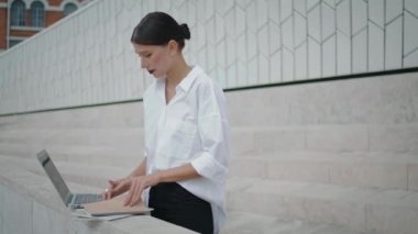 Young girl freelancer working laptop outdoors checking information in papers. Attractive elegant business woman searching data in documents standing street. Busy lady working remotely with notebook.