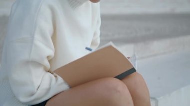 Unknown young woman writing into notebook sitting staircase close up. Girl hands with pen making notes in notepad diary outdoors. Unrecognizable freelancer working sitting on stairs. Urban lifestyle.