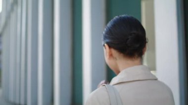 Back view of unknown walking woman straightening hairstyle close up. Unrecognizable young girl going near modern city building wearing stylish coat. Active pretty lady with hair bunch hurrying on work