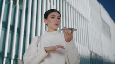 Happy stylish woman recording voice message on smartphone microphone standing street close up. Young lady talking in phone loudspeaker using mobile assistant app outdoors. Mobile technology concept.