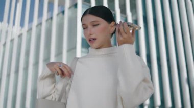 Stylish young girl talking in phone loudspeaker standing modern city street close up. Smart businesswoman recording voice message in smartphone microphone outdoors. Woman using mobile application.