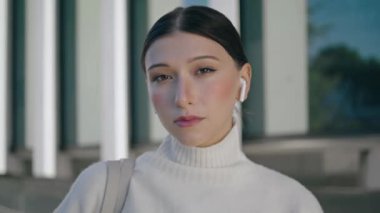 Serious gorgeous businesswoman looking camera confidently standing city street close up. Portrait of attractive young woman wearing modern wireless headphones. Beautiful girl model posing outdoors.