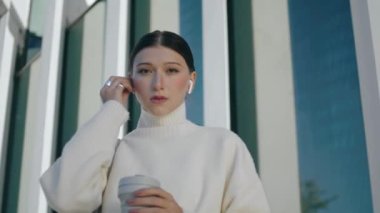 Kendine güvenen çekici iş kadını, şehir binasının yanında duran kulaklıklı, kablosuz kulaklık takıyor. Ciddi bir genç bayan, müzik dinlemek için kulaklık kullanıyor. Elinde kahve fincanı var. Modern teknoloji.