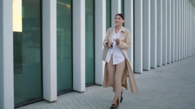 Cheerful walking girl talking mobile by wireless earbuds holding modern smartphone in hand. Stylish young business lady going on work speaking telephone. Smart woman using internet technology outdoors