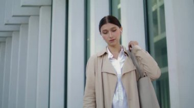 Attractive fashion model posing city street standing near modern building close up. Portrait of elegant woman wearing wireless white earbuds looking camera confidently. Beautiful businesswoman alone.