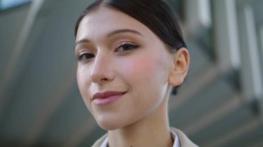 Portrait of successful business woman looking camera confidently with mysterious smile. Beautiful girl face with elegant hairstyle in front modern city building close up. Smiling lady posing outdoors.