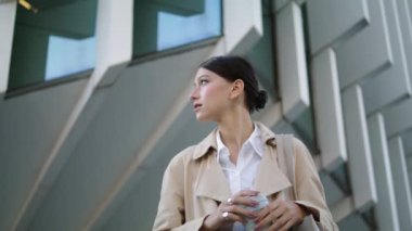 Young stylish woman standing city street with coffee takeaway looking around close up. Attractive millennial girl stopped near modern building with paper cup. Pretty lady drinking hot beverage.