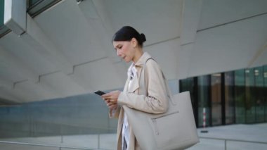 Serious young businesswoman messaging by smartphone outside wearing elegant coat. Worried attractive woman reading bad news looking phone screen. Anxious company manager standing street with mobile.