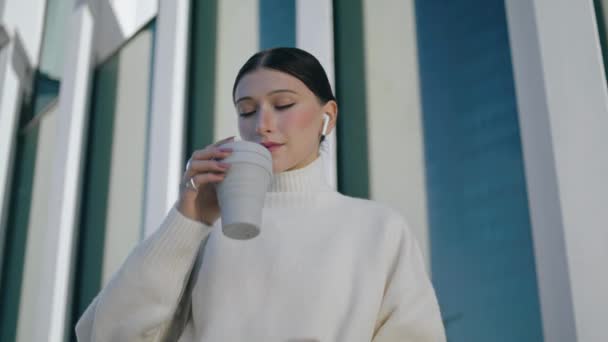 在近旁的现代城市建筑前 有一个悠闲的 时髦的女孩正在喝咖啡 迷人的年轻女子拿着纸杯品酒 漂亮的女士穿着耳塞喜欢工间休息 — 图库视频影像