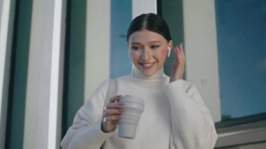Beautiful business woman talking wearing wireless earphones in front urban building closeup. Attractive confident lady speaking by headset. Young successful girl relaxing with cup coffee on work break