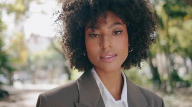 Güzel, mutlu egzotik bir kadının portresi doğanın üzerinde tek başına duruyor. Çekici Afro-Amerikan kadın yüzü. Yeşil ağaçların önünde doğal kıvırcık saçlı. Gülümseyen kadın saçlarına dokunuyor.