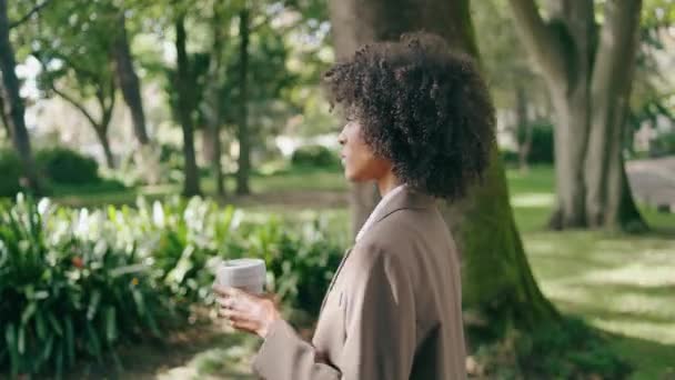 コーヒーペーパーカップを閉じて都会の公園を歩く忙しい女性を自信を持って 熱い飲み物を飲む仕事に急いで魅力的な若いアフリカ系アメリカ人のビジネス女性が持ち帰ります エレガントな女の子は路地に行く — ストック動画