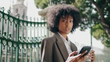 Successful beautiful woman scrolling phone holding paper cup with beverage takeaway outdoors closeup. Confident african american girl drinking coffee standing at iron gates. Stylish lady relax in park