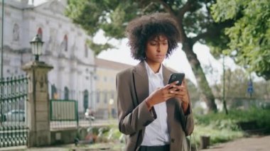 Focused woman surfing internet on smartphone standing alone at city park closeup. Attractive serious african american business lady typing phone message outdoor. Pretty girl using mobile on work break