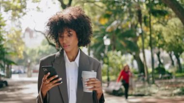 Confident businesswoman calling standing city park with paper coffee cup close up. Curly attractive african american girl using phone wearing elegant suit. Stylish woman holding mug takeaway drink.