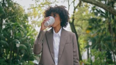 Young stylish business lady going at city park alley with paper cup hot coffee closeup. Confident african american woman holding drink takeaway walking alone. Curly girl in elegant suit looking phone.