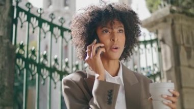 Confident gorgeous african american woman calling mobile holding coffee takeaway outdoors close up. Curly young business lady talking phone standing at iron gates. Stylish girl holding paper cup.