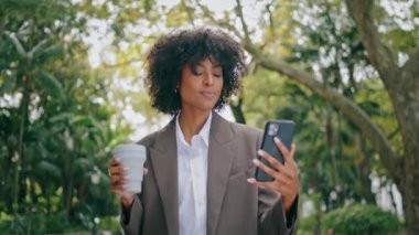 Relaxed woman talking phone video call walking city park hold paper coffee cup closeup. Attractive confident african american business lady speaking online outdoors. Pretty girl use smartphone camera