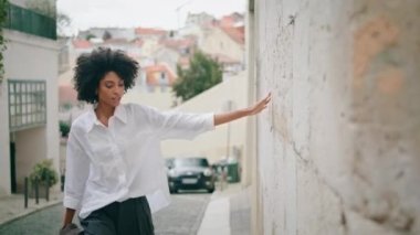 Relaxed stylish model walking alone watching city architecture wearing white shirt. Beautiful african american woman going street touching old building wall. Curly tourist enjoying cityscape outdoors.