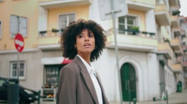 Carefree gorgeous girl looking around standing at cozy city street alone close up. Attractive african american woman exploring town district wearing elegant jacket. Curly lady smiling enjoy weekend.