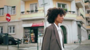 Confident curly businesswoman crossing road summer day close up. Awesome african american woman walking crosswalk beautiful town. Young stylish lady going on modern city street looking camera.