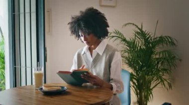 Curly young girl turning book page sitting at wooden table cozy cafe alone. Attractive african american woman reading interesting literature at breakfast in cafeteria. Relaxed brunette enjoying novel.