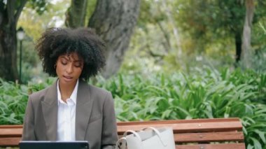 Relaxed smiling girl sitting park bench with laptop close up. Attractive african american woman chatting using modern notebook outdoors. Stylish businesswoman wearing suit making remote work on nature