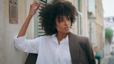 Attractive confident business woman standing leaning on wall wearing stylish jacket close up. Relaxed african american girl posing touching lush curly hairstyle. Young beautiful lady relaxing outdoors