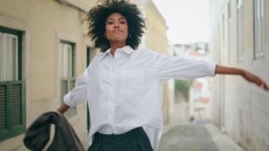 Happy cheerful woman tourist walking city street raising hands with jacket rejoicing beautiful day. Curly african american girl relaxing going near old town buildings. Attractive lady smiling close up