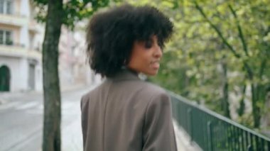 Carefree relaxed business woman walking city street wearing elegant jacket close up. Back view of attractive african american girl going near town road. Curly cute young lady relaxing after work day.