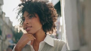 Güzel, çekici bir bayan güneşli şehir caddesinde rüya görüyor. Yaklaş. Mutlu Afro-Amerikan kadın dışarıda, parlak güneş ışığı gören kameranın altında duruyor. Kasabadaki güzel kıvırcık esmerin portresi..