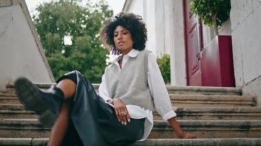 Elegant young woman posing on staircase looking camera confidently close up. Curly confident african american girl sitting on stairs wearing stylish outfit. Attractive brunette crossing sexy legs.