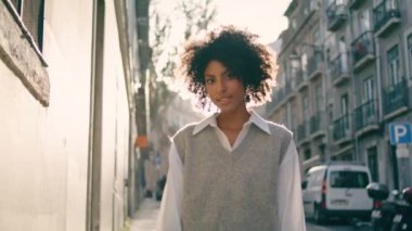 Cute attractive girl smiling walking city street close up. Beautiful african american woman with curly hairstyle looking camera posing outdoors. Charming happy lady brunette enjoying town at weekend.