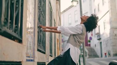 Stylish fashion model posing street holding on bars old building window. Curly african american woman relaxing on cozy town street. Attractive carefree girl enjoy calm urban walk at summer weekend.