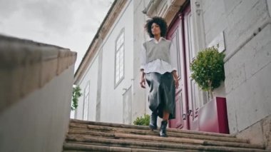 Stylish young girl walking down stairs cozy city cloudy day. Curly attractive african american woman enjoy calm stroll on staircase alone. Active fashionable brunette going downstairs exploring town.