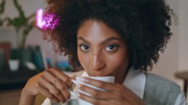 Smiling young woman drinking fresh tasty latte looking camera close up. Portrait of happy attractive girl sitting cafe with white cup aromatic coffee. Curly relaxed african american lady enjoy drink.