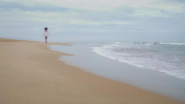 在阴郁的一天 一个身材苗条的女孩在海滨散步 松懈的非洲裔美国女人在平静的大海中休息 浪漫的游客穿着白色比基尼去沙滩上的海岸线 思考个人问题的忧心忡忡者 — 图库视频影像