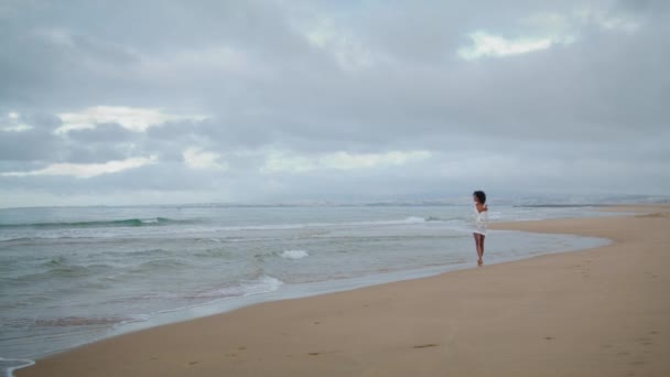 梦幻般的女人在多云的日子走在海滩上 浪漫的非洲裔美国女孩穿着白衣赤脚享受着大海的海岸线 迷人的黑发女士放松地凝视着岸边的波浪 — 图库视频影像