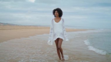 Dreamy woman walking coastline in white outfit. Thoughtful curly girl going sand beach on gloomy day alone. Worried african american contemplating personal problems at sea. Loneliness crisis concept.