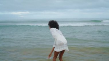 Smiling girl enjoying ocean waves in bikini. Happy woman having fun at sea shore alone. Joyful slim african american going throwing water on cloudy day. Satisfied curly lady relax on seaside vacation.
