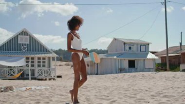 Plaj kızı deniz kenarındaki evlerde tenis raketi tutuyor. Sıska seksi bikinili kadın, hafta sonu bilinmeyen partneriyle oyun oynarken eğleniyor. Afro-amerikalı oyuncu seyahat kıyısı aktivitelerini seviyor.